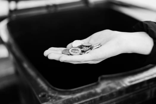 A kid girl hand throws euro coins into a trash can dumpster, street shot.