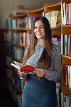 Smiling girl with a book in her hands standing near bookshelves. looking to the camera
