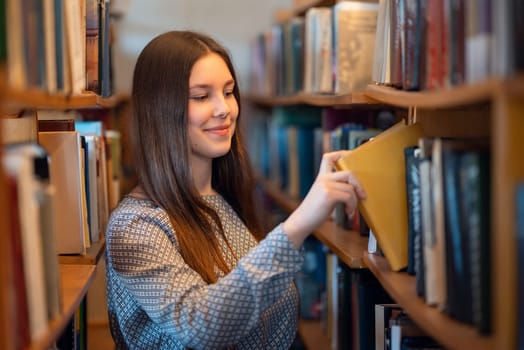 Young smiling girl standing near bookshelves in library taking books for reading, literature lesson