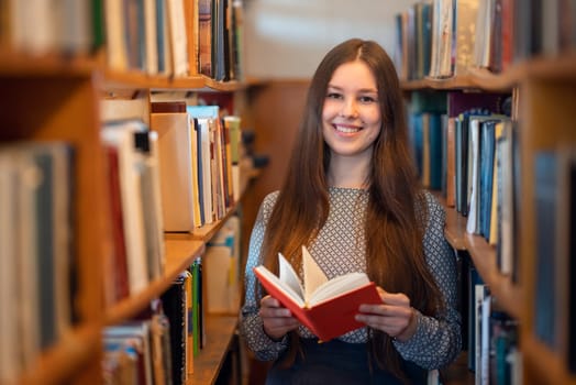 Cheerful brunette student standing with a book in her hands between the rows of bookshelves in library