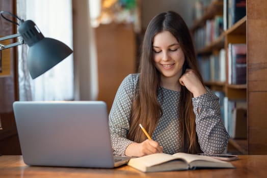 Girl sitting at the desk, writing notes, preparing for classes, using laptop