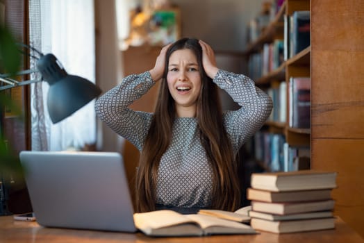 Woman sitting at a table full of books and textbooks, putting the hands on her head in despair