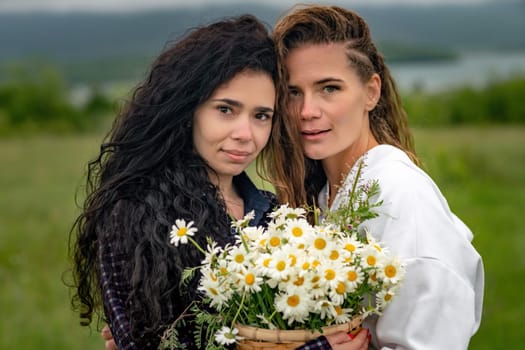 Two women enjoy nature in a field of daisies. Girlfriends hugging hold a bouquet of daisies and look at the camera