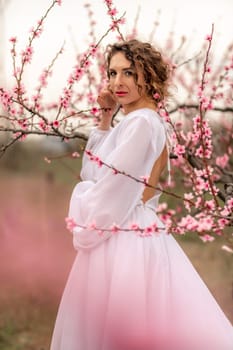 Woman peach blossom. Happy curly woman in white dress walking in the garden of blossoming peach trees in spring.