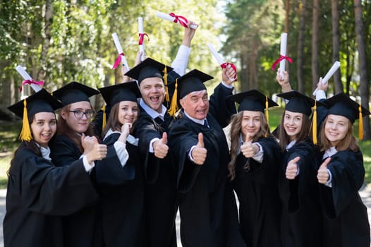 A group of graduates in robes give a thumbs up outdoors. Elderly student