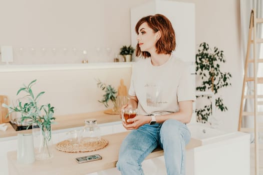 Woman kitchen tea. Carefree woman rests in a cozy kitchen, drinks tea, sits on the table and dreams, free space. Woman holding cup while enjoying hot drink at home.