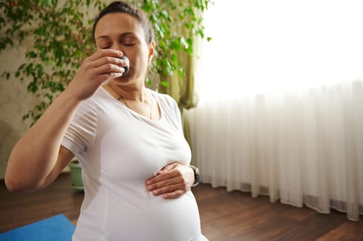 African American pretty pregnant woman holding her belly, enjoying tea ceremony after prenatal fitness exercises at home. Gravid expectant mother drinks puer tea, relaxes after pregnancy yoga practice