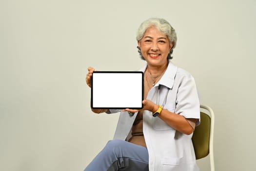 Portrait of senior female doctor showing digital tablet with blank mockup screen for medical healthcare tech website advertise.