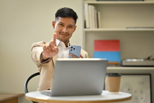 Image of smiling male entrepreneur holding smartphone, showing and pointing up with fingers number one.