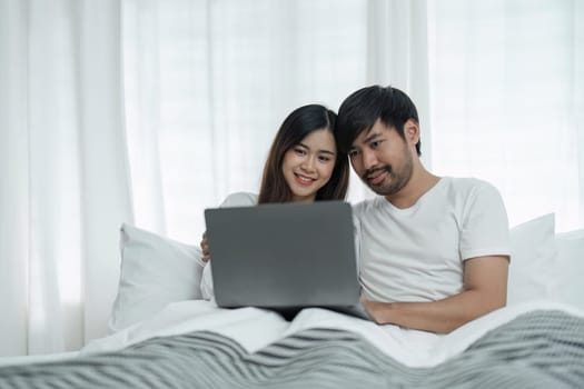 Young couple using laptop sitting on the bed at home. Full length of couple watching movie on laptop. Happy man and woman are relaxing on bed. They are spending leisure time at home.