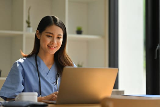 Smiling female doctor reading electronic medical records on laptop computer while sitting at modern workplace.