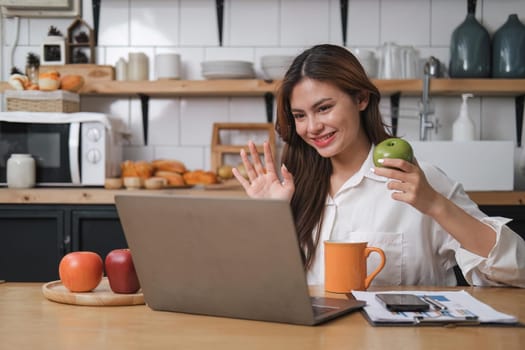smiling young woman on a video call with colleagues, sitting on a kitchen with laptop computer, waving.