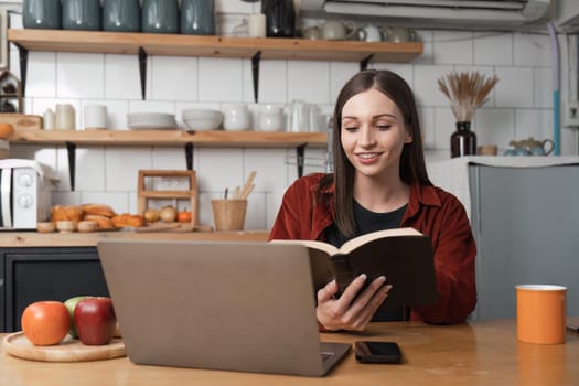 Beautiful woman sitting at table reading book in nice modern kitchen at home, she is positive and smiling.