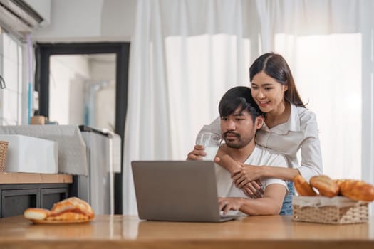 Handsome man sitting near his wife at kitchen. Family couple see social media, surf the web while sitting at kitchen table with generic laptop. Couple working with laptop at home.