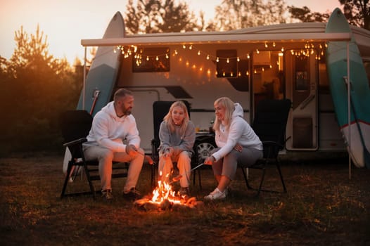 A family cooks sausages on a bonfire near their motorhome in the woods.