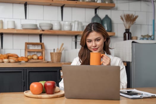 Young woman drinking morning coffee and looking at monitor of laptop while sitting in the kitchen at home.