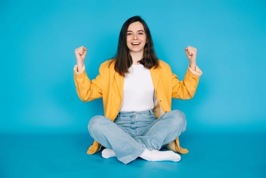 Vibrant Portrait of a Joyful Girl Celebrating Success with Raised Fists and a Victorious 'Yes ' - Isolated on Blue Background - Positive, Optimistic Emotions.