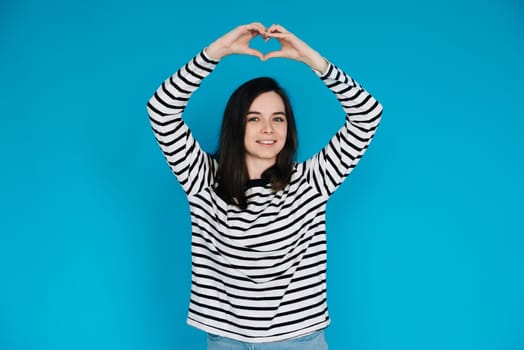 Happy Woman in Striped Sweater Spreading Love and Positivity - Smiling Girl with Raised Arms Forming Heart Shape, Expressing Joy and Affection - Isolated Blue Background - Ideal for Happiness, Love,