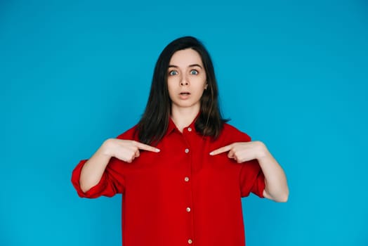 A photo of a lovely impressed young woman in a red shirt, surprised face, pointing her finger, isolated on a blue background - Perfect for Emphasizing Surprise and Amazement.