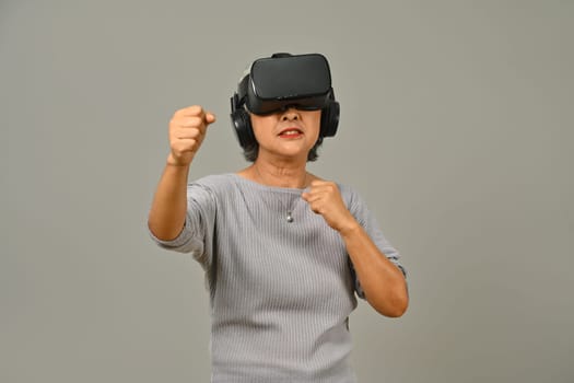 Happy senior woman in boxing stance playing action simulator game, fighting with fist in VR headset. Modern technologies, innovations.