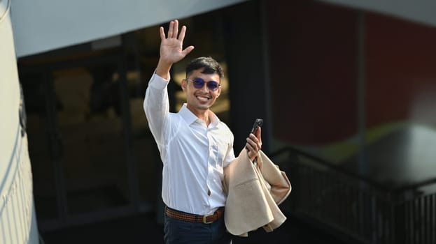 Friendly businessman in white shirt waving hand, looking at camera with toothy smile, greeting someone.