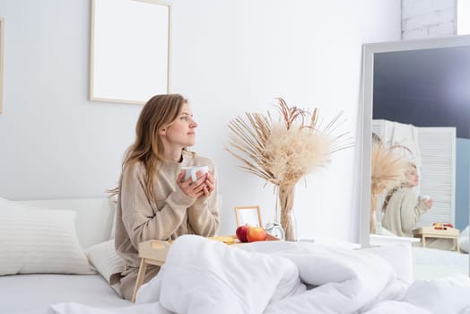 caucasian woman waking up in her bed in the morning. young millennial woman sitting on the bed in the morning drinking coffee, light and airy room