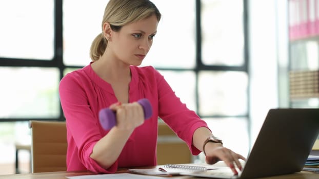 Businesswoman lifting dumbbell and working on laptop in office. Feminism and success of women in business concept