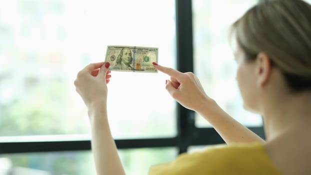 Woman studying watermarks on money dollars in front of window closeup. Counterfeit money concept