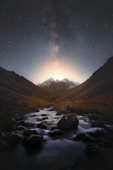 Milky Way over snowy mountains and mountain river at night. Landscape with high cliffs covered with snow, blue and purple starry sky, reflection in the water. Sky with stars. Bright milky way, space.