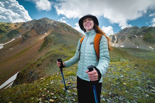 Portrait, the girl is happy and smiling while hiking in the mountains. Beautiful young caucasian sporty woman joyful stands with a backpack and looks at the camera.
