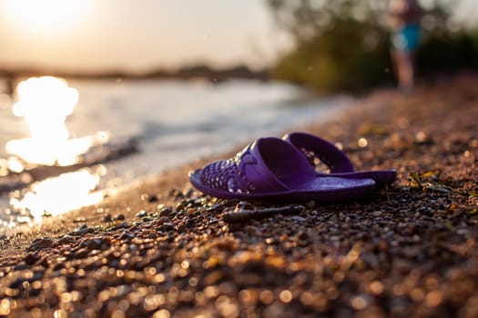 Close-up of rubber slippers on the shore of a lake or river made of small stones against a colorful sunset background. A beautiful place in nature for family holidays and swimming.