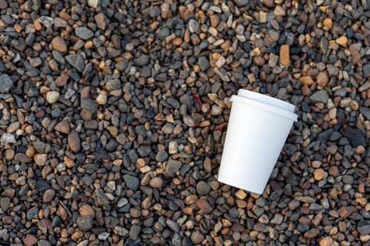 A white paper cup of coffee or tea lies on the rocky shore. A mug of hot drink on a pebble. Beautiful rocky background. free copy space.