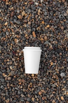 A white paper cup of coffee or tea lies on the rocky shore. A mug of hot drink on a pebble. Beautiful rocky background. free copy space.