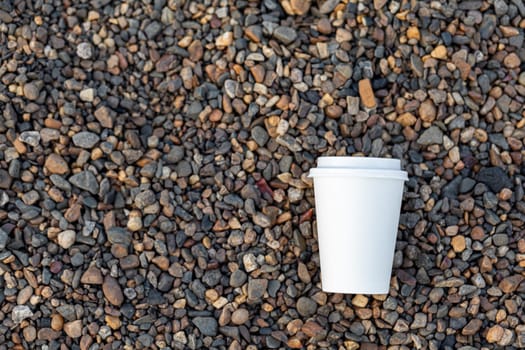 A white paper cup of coffee or tea lies on the rocky shore. A mug of hot drink on a pebble. Beautiful rocky background. free copy space.