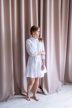 Beautiful caucasian woman in white summer fashion dress standing barefoot on beige curtain background, looking away. Pastel colors