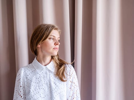 Beautiful caucasian woman in white summer fashion dress standing on beige curtain background, looking away. Pastel colors