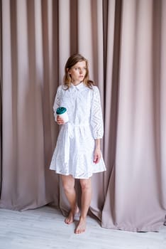 Beautiful caucasian woman in white summer fashion dress standing barefoot on beige curtain background, looking away. Pastel colors