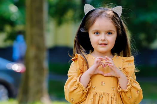 Portrait of a cute little girl in the park and shows a heart sign with her hands.