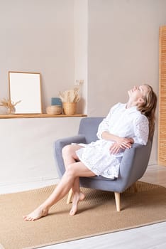 Beautiful caucasian woman in white summer fashion dress sitting on soft chair barefoot, looking away. Boho or scandi interior