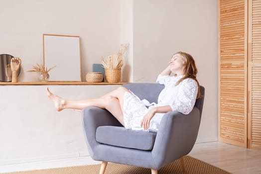 Beautiful caucasian woman in white summer fashion dress sitting on soft chair barefoot, looking away. Boho or scandi interior