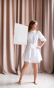 barefoot woman in beautiful white dress holding blank frame, beige curtain background, mockup design