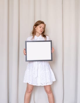 barefoot woman in beautiful white dress holding blank frame, beige curtain background, mockup design