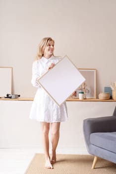 barefoot woman in beautiful white dress holding blank frame in living room, mockup design