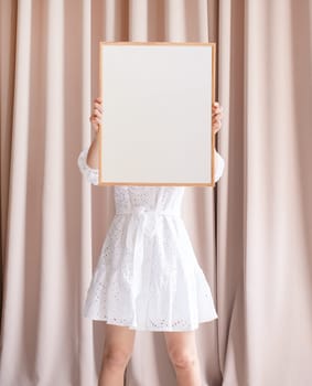 barefoot woman in beautiful white dress holding blank frame in front of her face, beige curtain background, mockup design