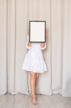 barefoot woman in beautiful white dress holding blank frame in front of her face, beige curtain background, mockup design