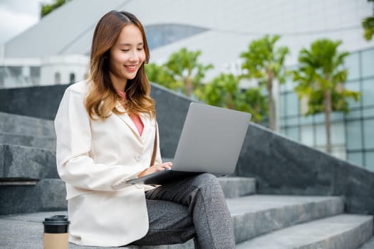 Happy young Asian woman freelance looking laptop with smile while working with coffee cup while sitting on stairs outdoors in the city. Remote working concept in the office.