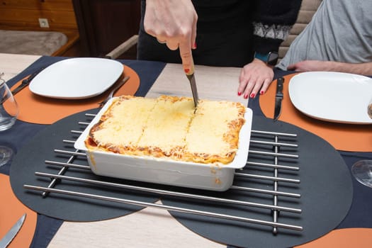 Woman cut with knife hot tasty home baking lasagna in ceramic casserole dish, whole family sit at the table, High quality photo