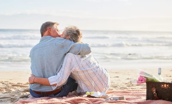 Senior couple, beach picnic and kiss with hug, romance and happiness in summer for anniversary celebration. Elderly man, old woman and basket for food, wine and outdoor for sunshine, waves and love.
