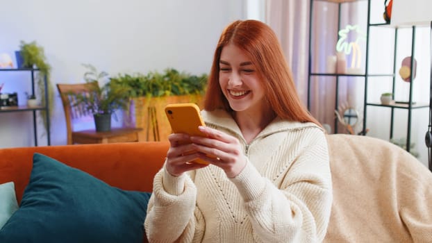 Happy young woman use mobile smartphone typing browsing say Wow yes found out great big win good news celebrate lottery jackpot doing winner gesture. Redhead girl at home in room sitting on sofa