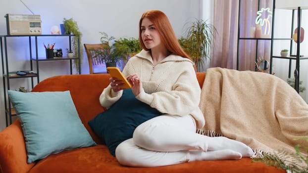 Redhead young woman sitting on sofa uses mobile phone smiling at home living room apartment. Girl texting share messages content on smartphone social media applications online watching relax movie